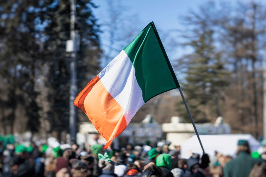 A close up shot of the Irish flag with blurry trees in the background up against a blue sky.